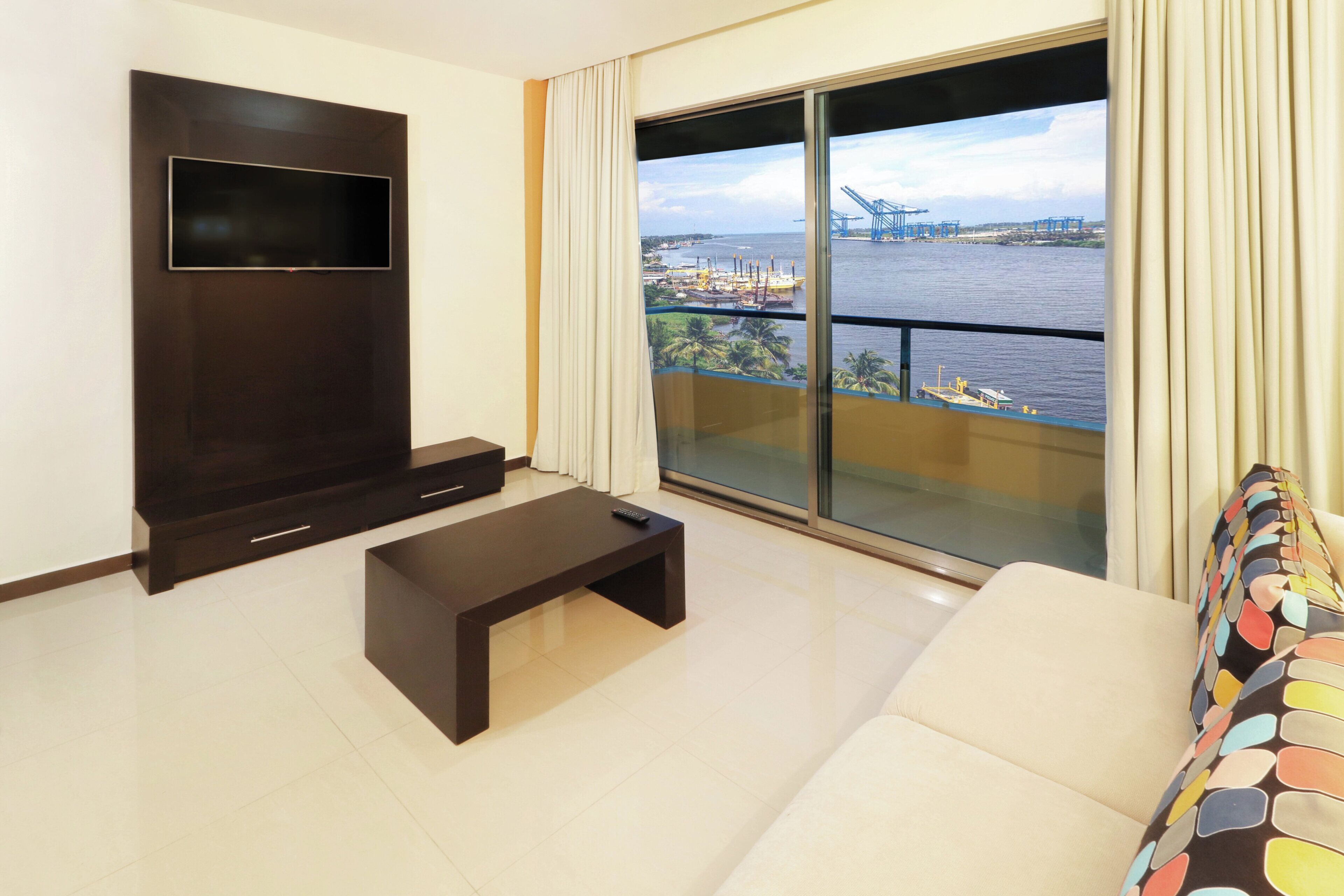 Un salon moderne avec un téléviseur à écran plat, une petite table basse et un canapé. Une grande fenêtre offre une vue sur un port avec des navires et des grues.