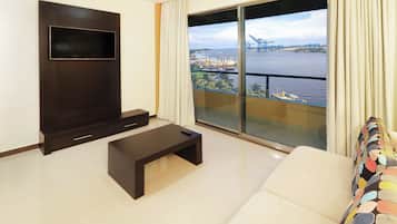 Un salon moderne avec un téléviseur à écran plat, une petite table basse et un canapé. Une grande fenêtre offre une vue sur un port avec des navires et des grues.