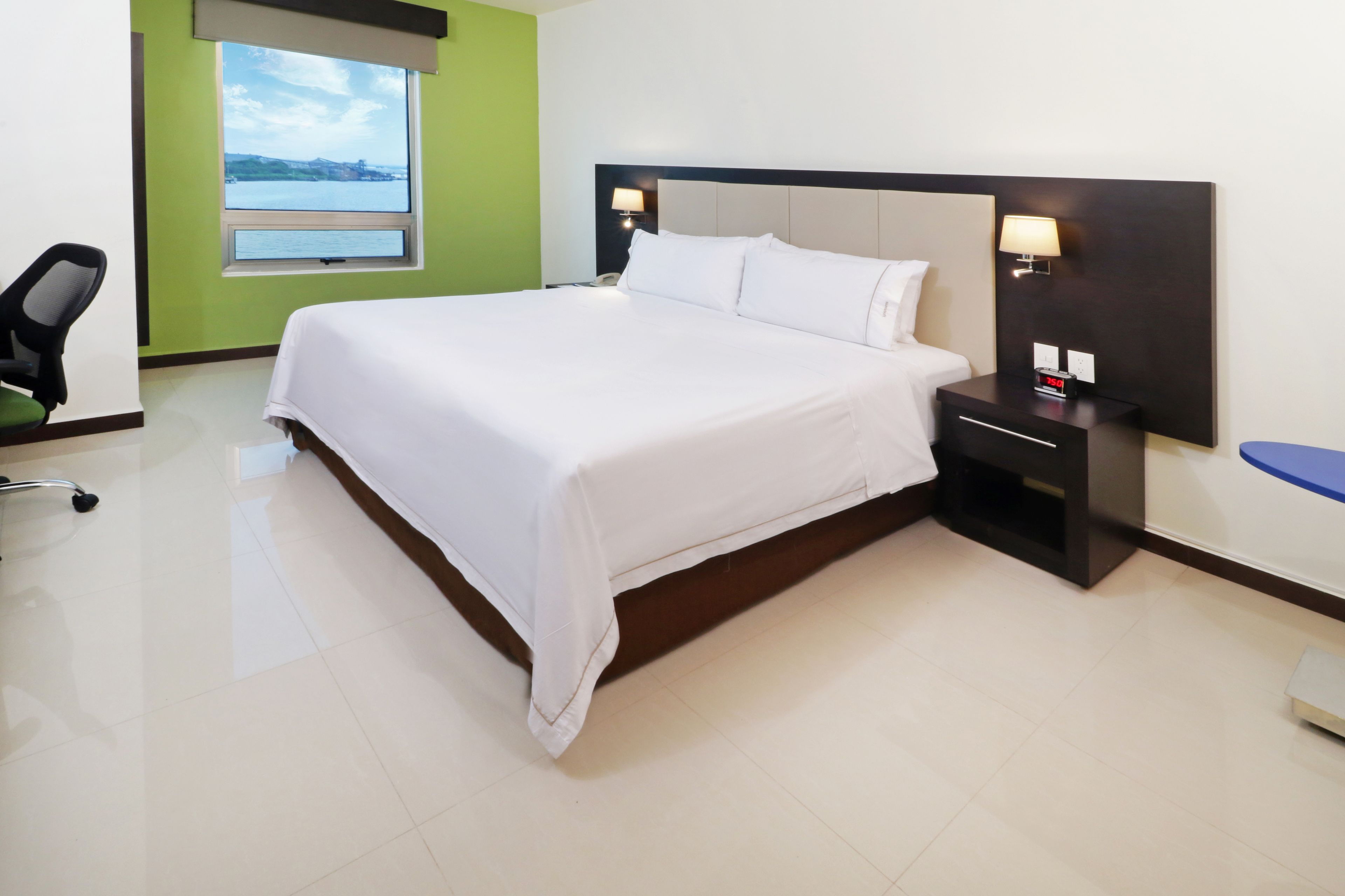 Habitación de hotel con una cama grande, un escritorio, una silla, una mesita de noche con lámpara y vistas al exterior.