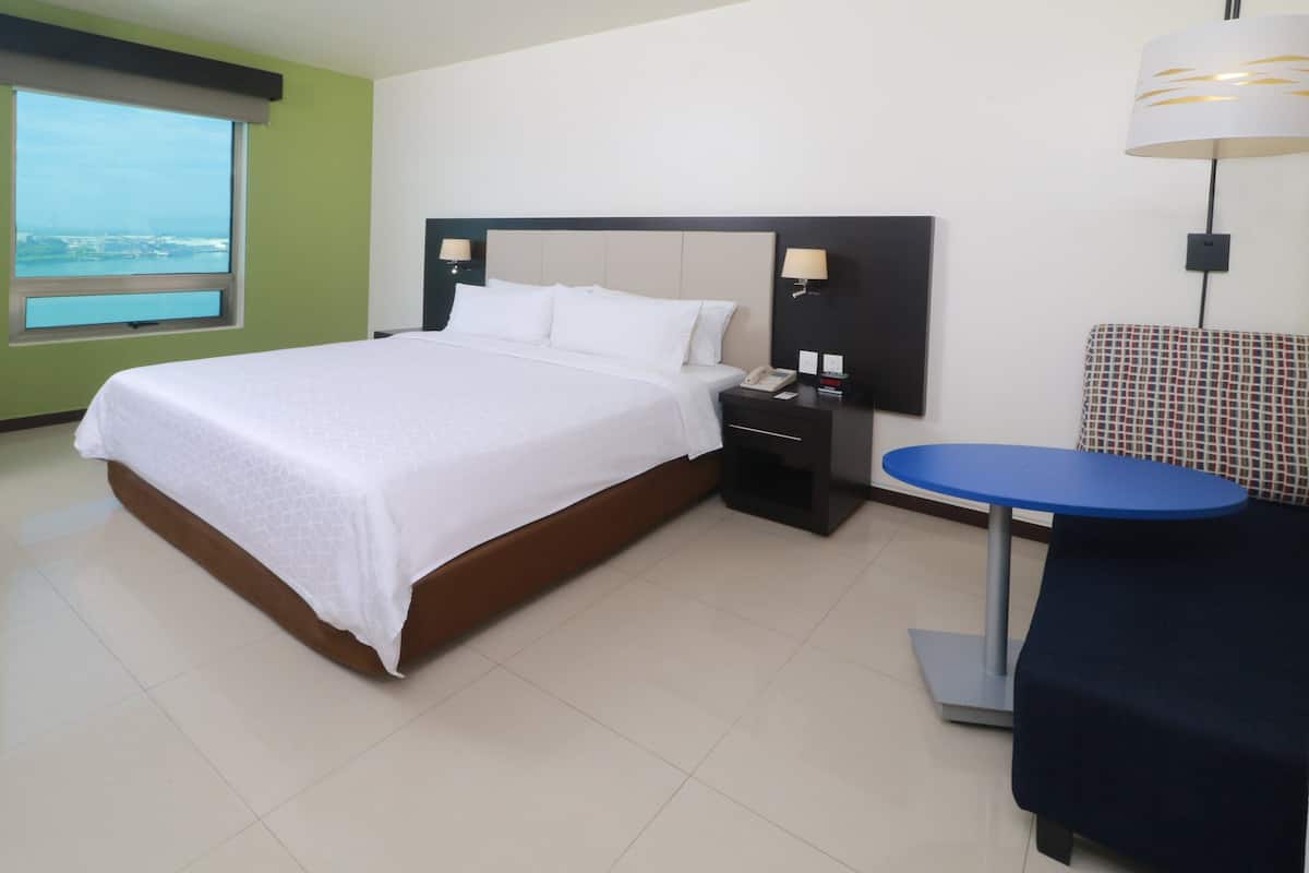 Quarto de hotel com uma cama grande, uma mesa de cabeceira com abajur, uma pequena mesa redonda e vista para a cidade pela janela.
