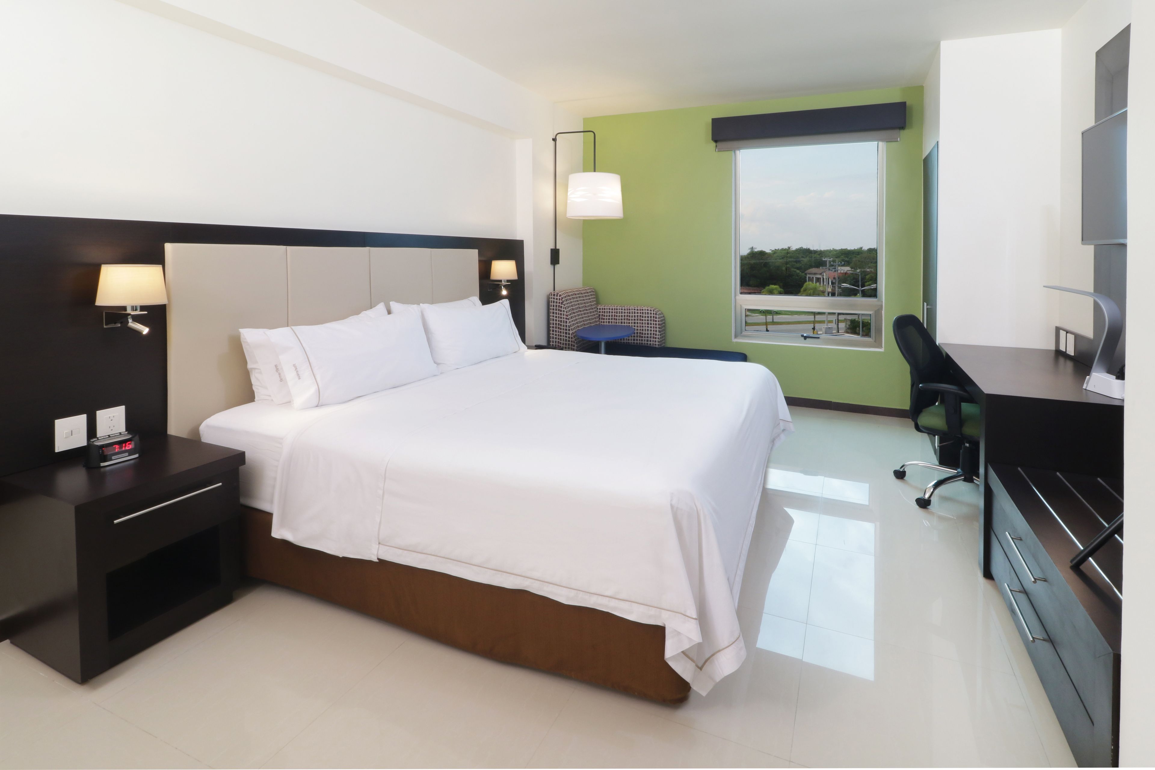A modern hotel room with a large bed, a desk, a chair, and a view of the city.