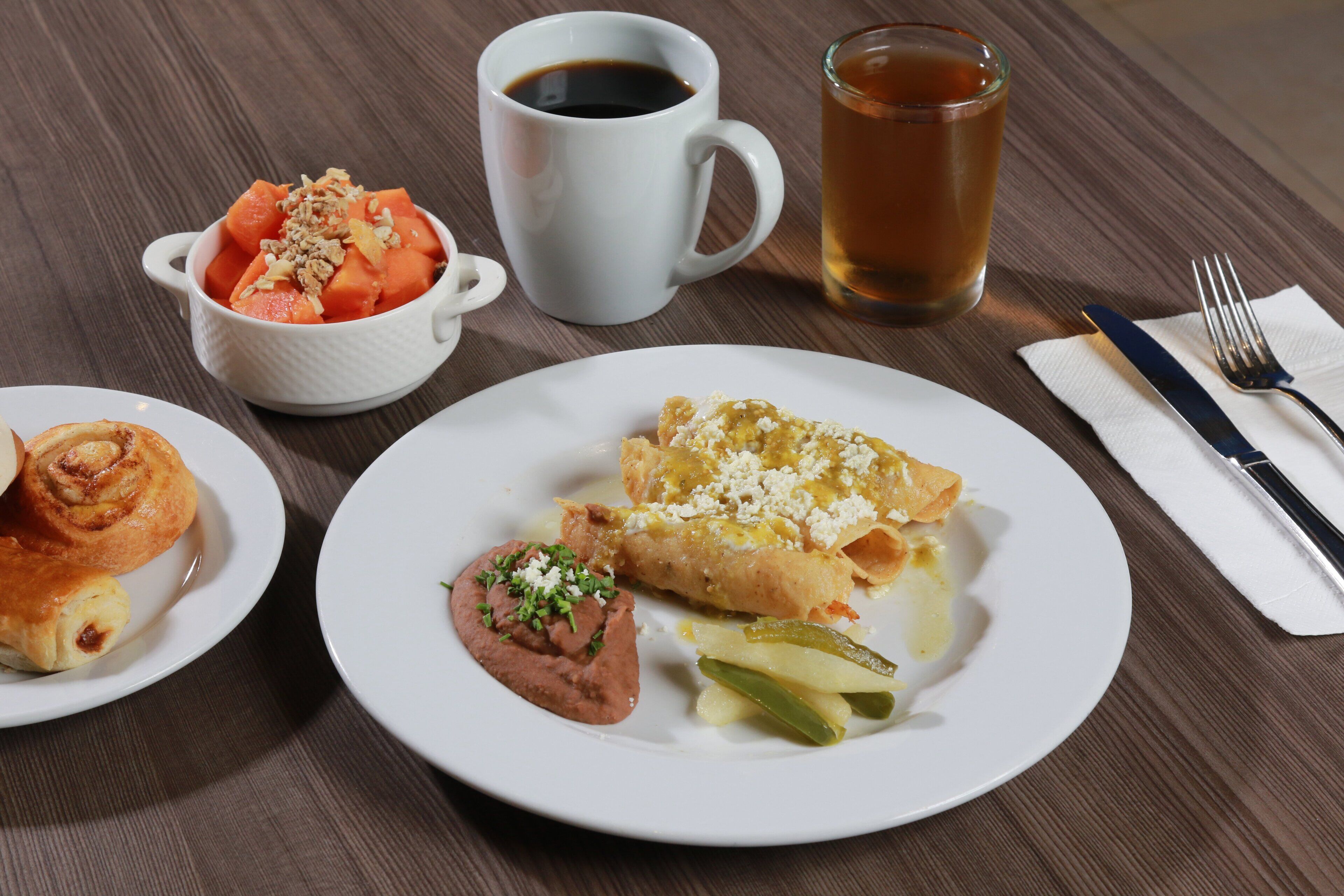 A table with a meal consisting of a plate with a rolled pastry, a bowl of fruit, a plate with a dip and pickles, a cup of coffee, and a glass of tea.