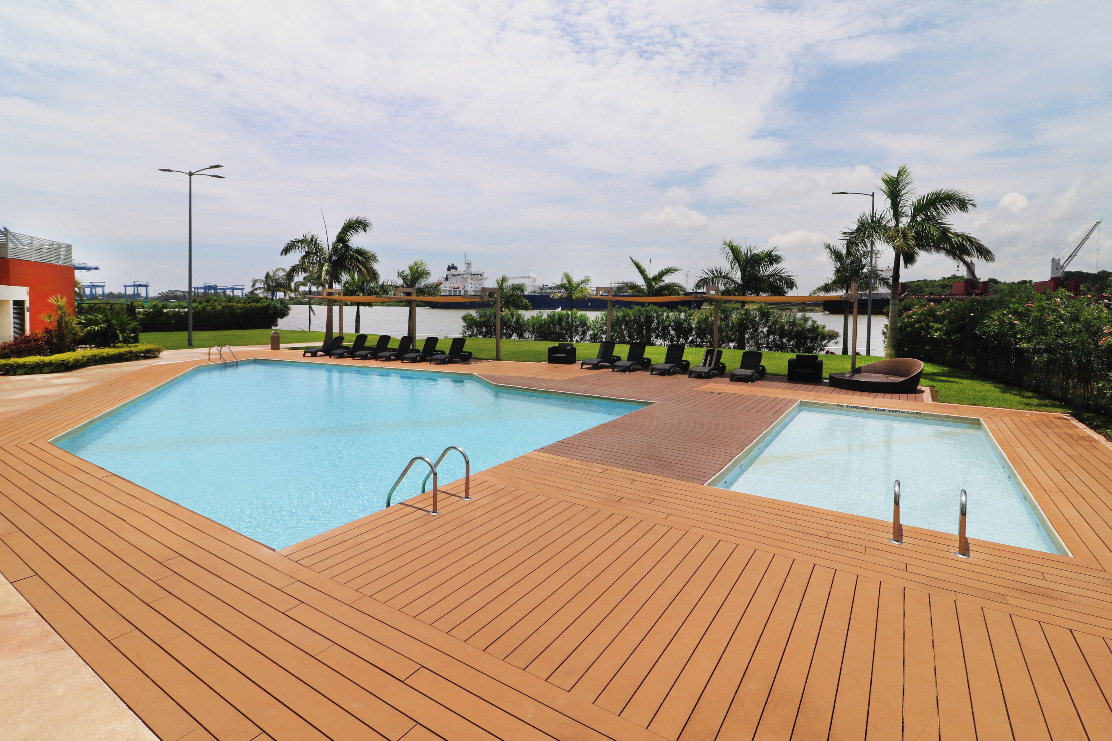 A rectangular swimming pool with a wooden deck, surrounded by lounge chairs and palm trees.