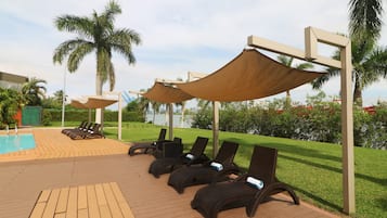 Zona junto a la piscina con tumbonas y sombrillas, rodeada de palmeras y un césped.