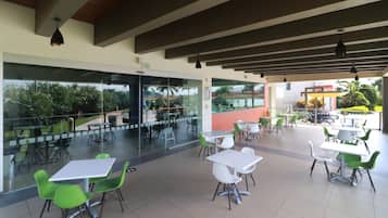Un café al aire libre con mesas y sillas, amplios ventanales y vistas al exterior.