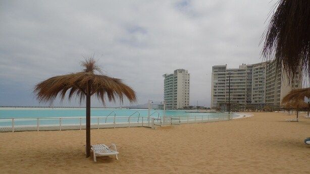 A beach with a thatched umbrella, a white lounge chair, and a pool area. - Alojamientos La Serena (La Serena)