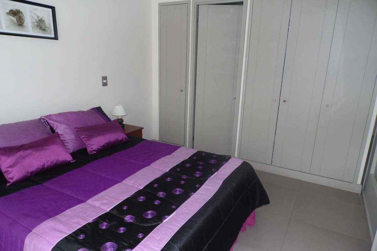 Une chambre à coucher avec un lit, une literie violette, une table de chevet, une lampe et une armoire.
