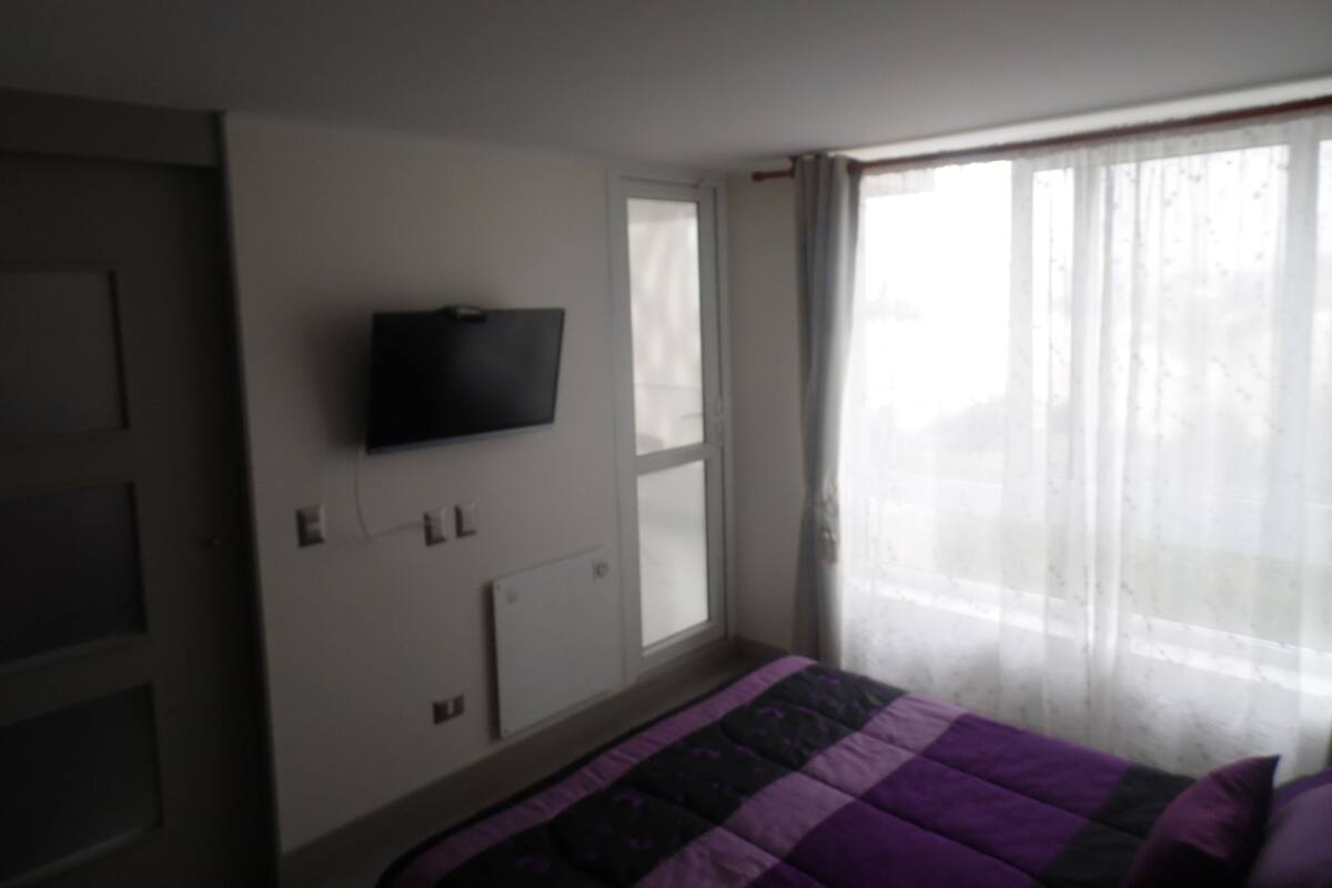 Une chambre à coucher avec un lit, un téléviseur fixé au mur et une fenêtre avec des rideaux.