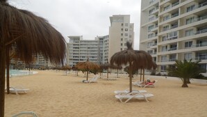 A beach with white lounge chairs and thatched umbrellas, surrounded by high-rise buildings. - Alojamientos La Serena (La Serena)
