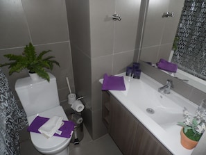 Un baño moderno con inodoro, lavamanos y espejo. Hay una planta sobre el inodoro, un rollo de papel higiénico y una toalla morada en el lavamanos.