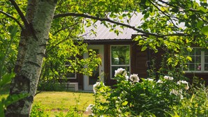 A wooden cabin with a large window and a door, surrounded by lush greenery and white flowers.