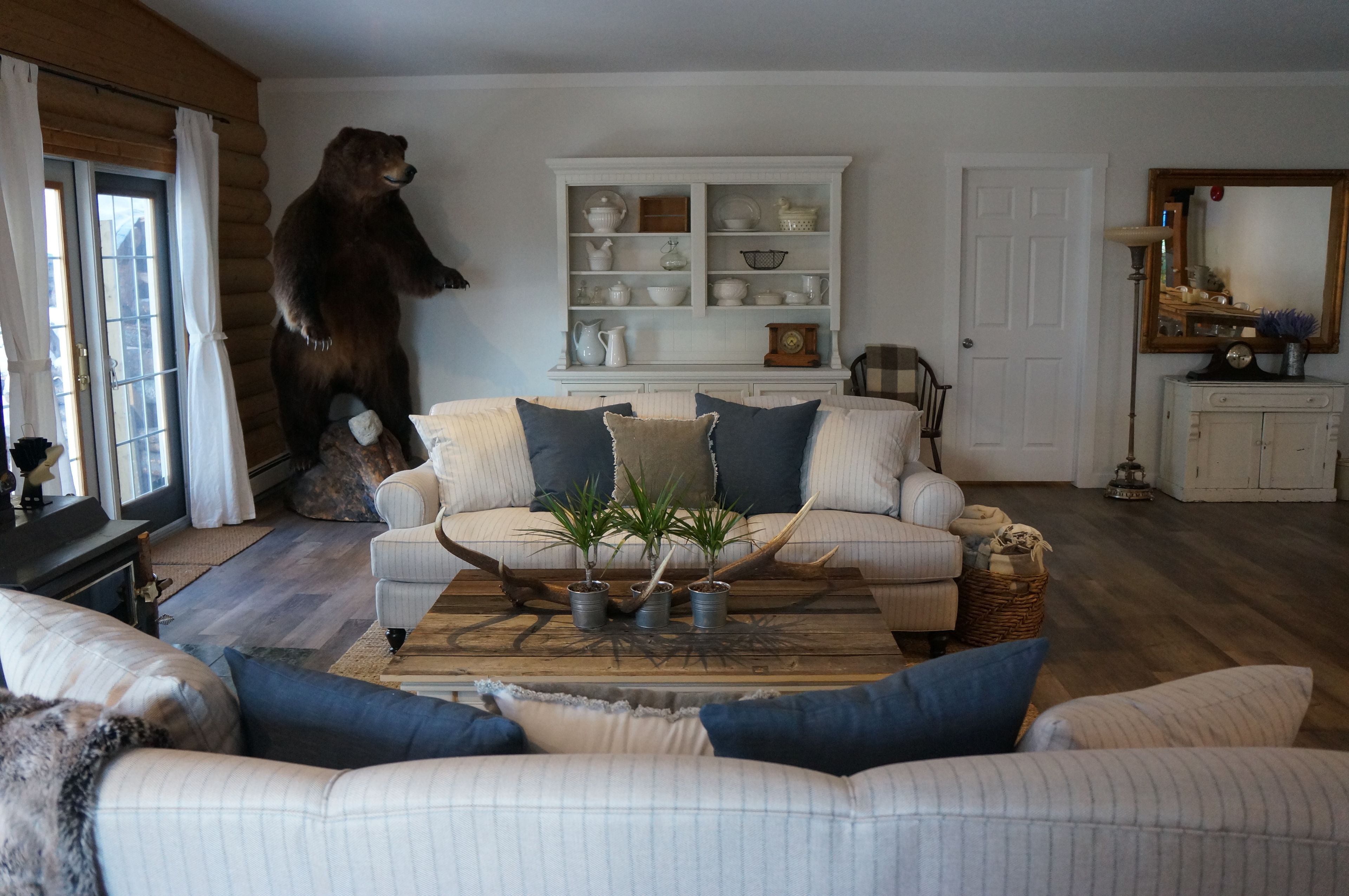 A living room with a sofa, a coffee table with plants, a bear statue, and a white cabinet with dishes.