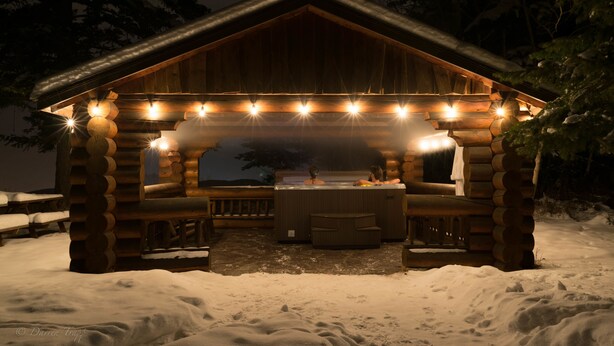 A wooden cabin with a hot tub and seating area, surrounded by snow.
