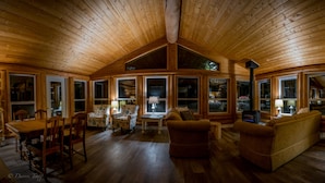 A cozy cabin interior with a wooden ceiling, large windows, a dining area, and a living room with sofas and a TV.