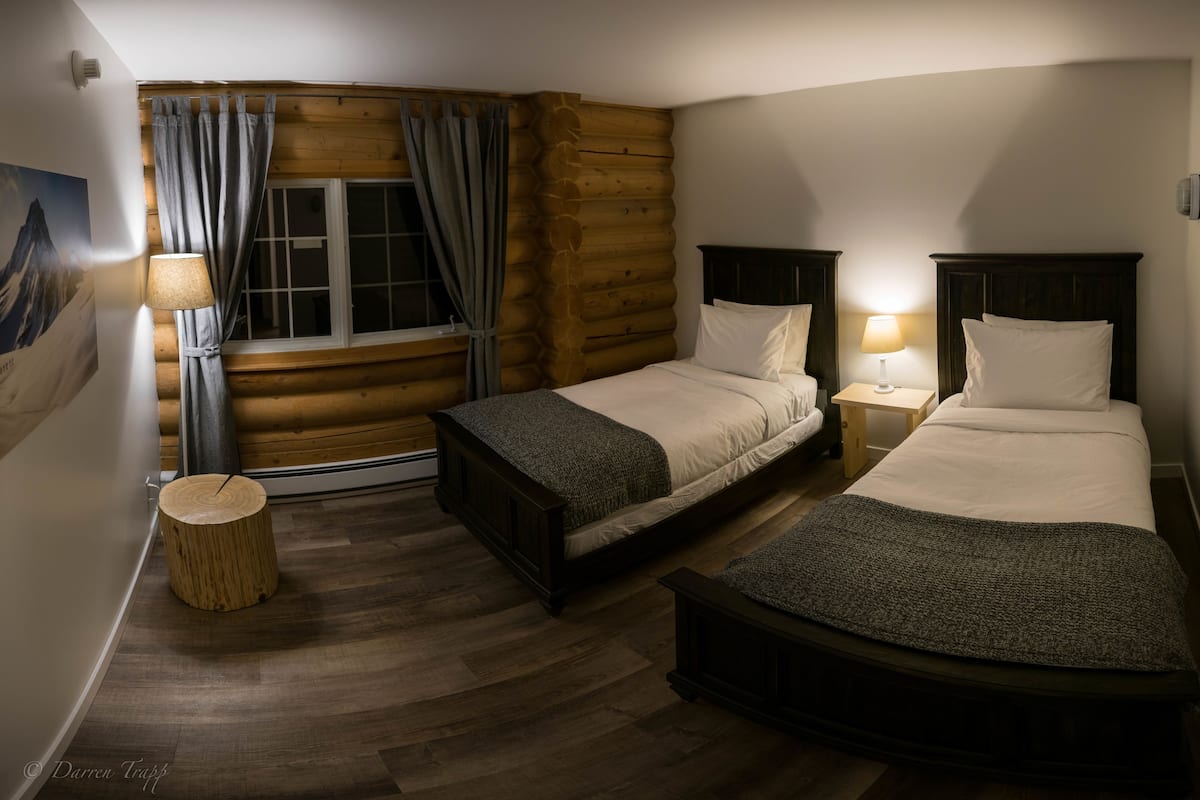 Un ambient acollidor a una cabana de fusta, amb dos llits, un terra de fusta i una finestra amb cortines.