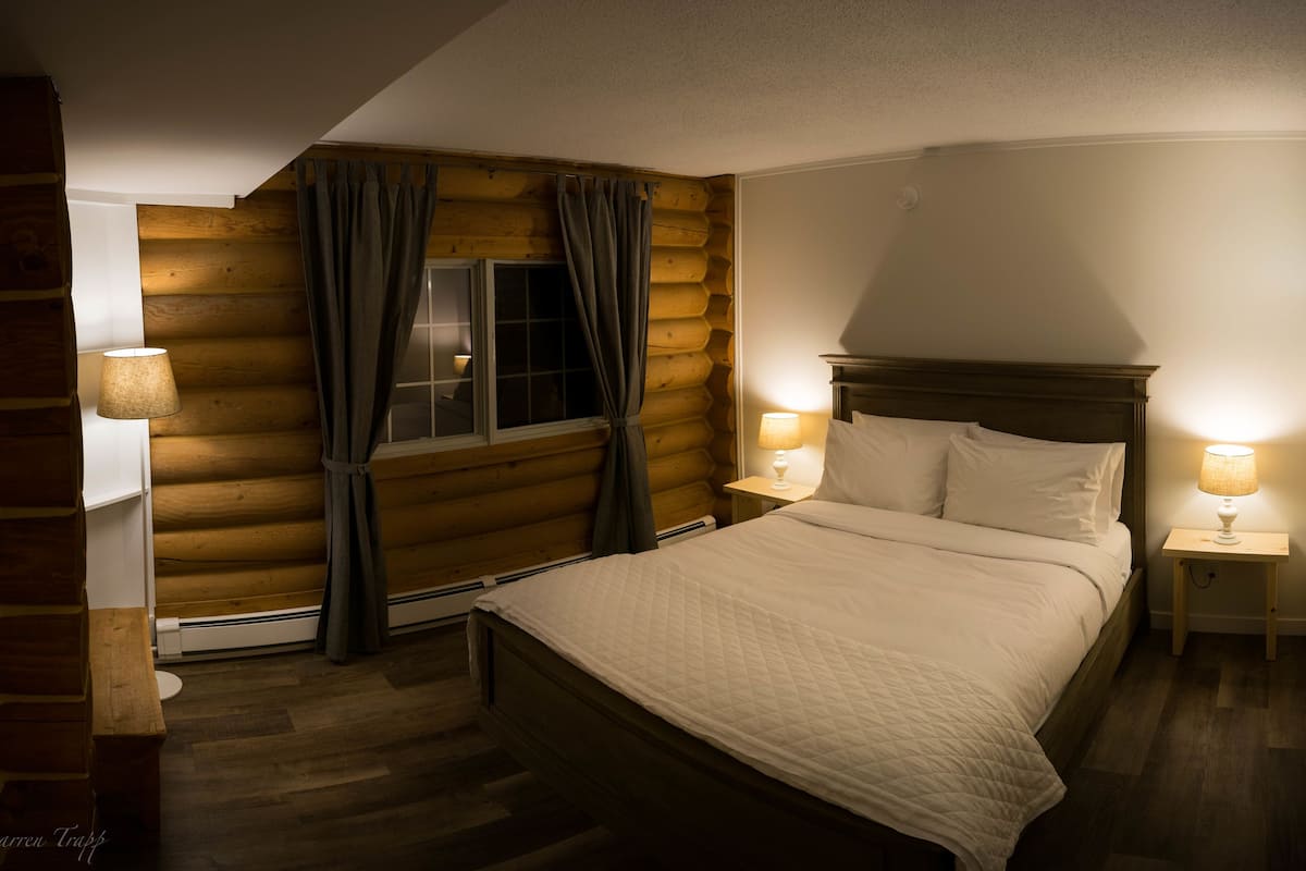 Un dormitori d'una cabana de fusta acollidora, amb un llit de fusta, dues làmpades de nit i una finestra amb cortines.