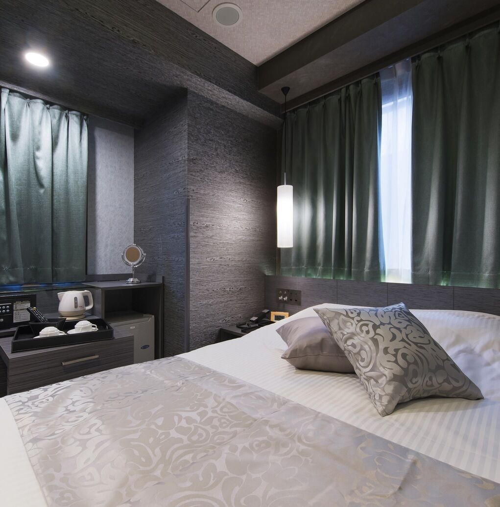 Una habitación de hotel moderna con cama, mesita de noche, espejo y vistas a la ciudad a través de amplias ventanas.