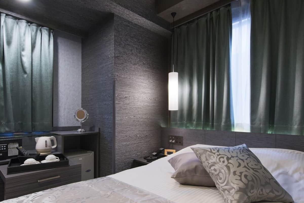 A modern hotel room with a bed, bedside table, mirror, and a view of the city through large windows.