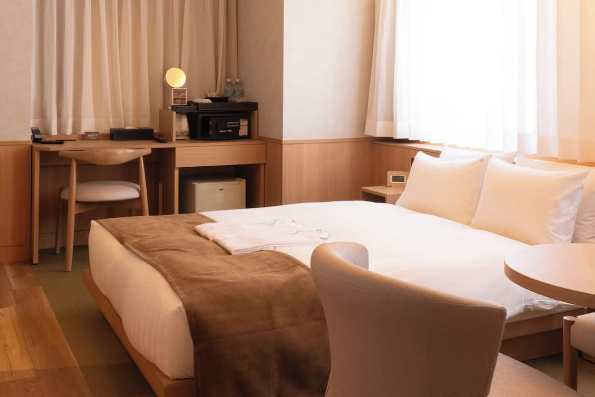 A hotel room with a bed, a desk, a chair, a bedside table, and a lamp.