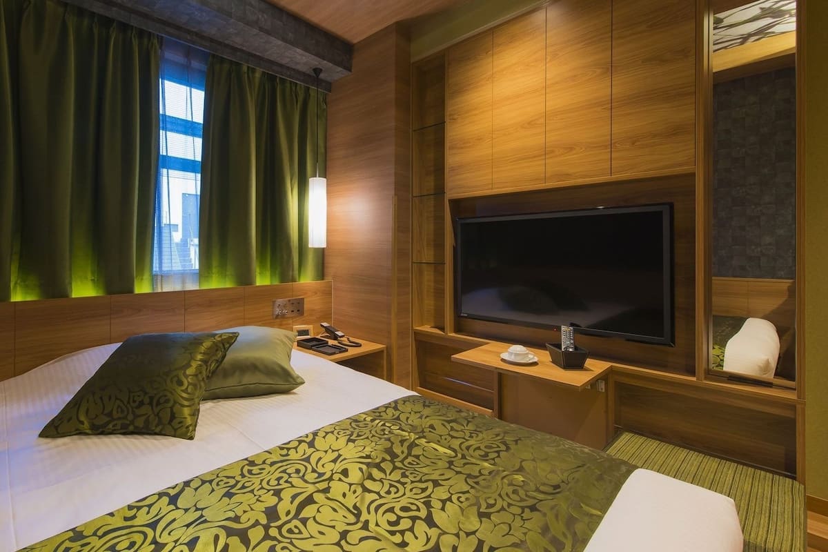A hotel room with a large bed, a television, a bedside table, and a window with green curtains.