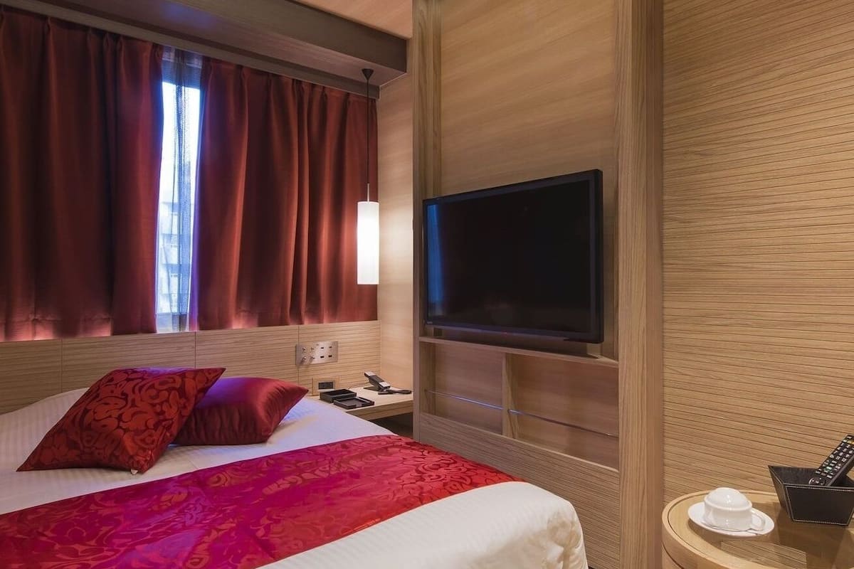 A hotel room with a bed, a television, a bedside table with a cup and remote, and a window with curtains.