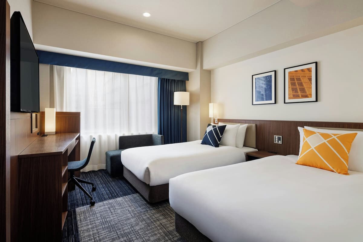 Une chambre d’hôtel avec deux lits, un bureau, une chaise et un téléviseur. Deux tableaux encadrés sont accrochés au mur.