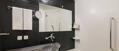 A modern bathroom with a black and white tiled wall, a white sink, a mirror, a shower, and a handrail.