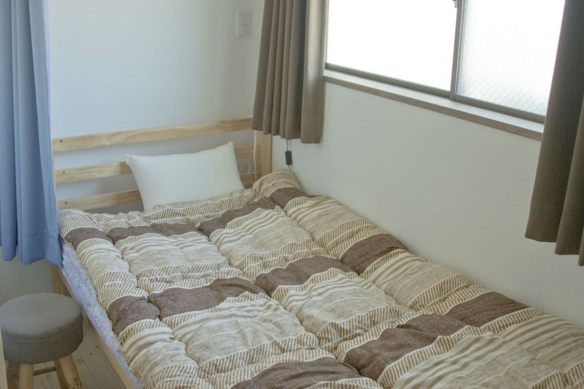 A single bed with a striped bedspread, a wooden headboard, a window with curtains, and a small stool.