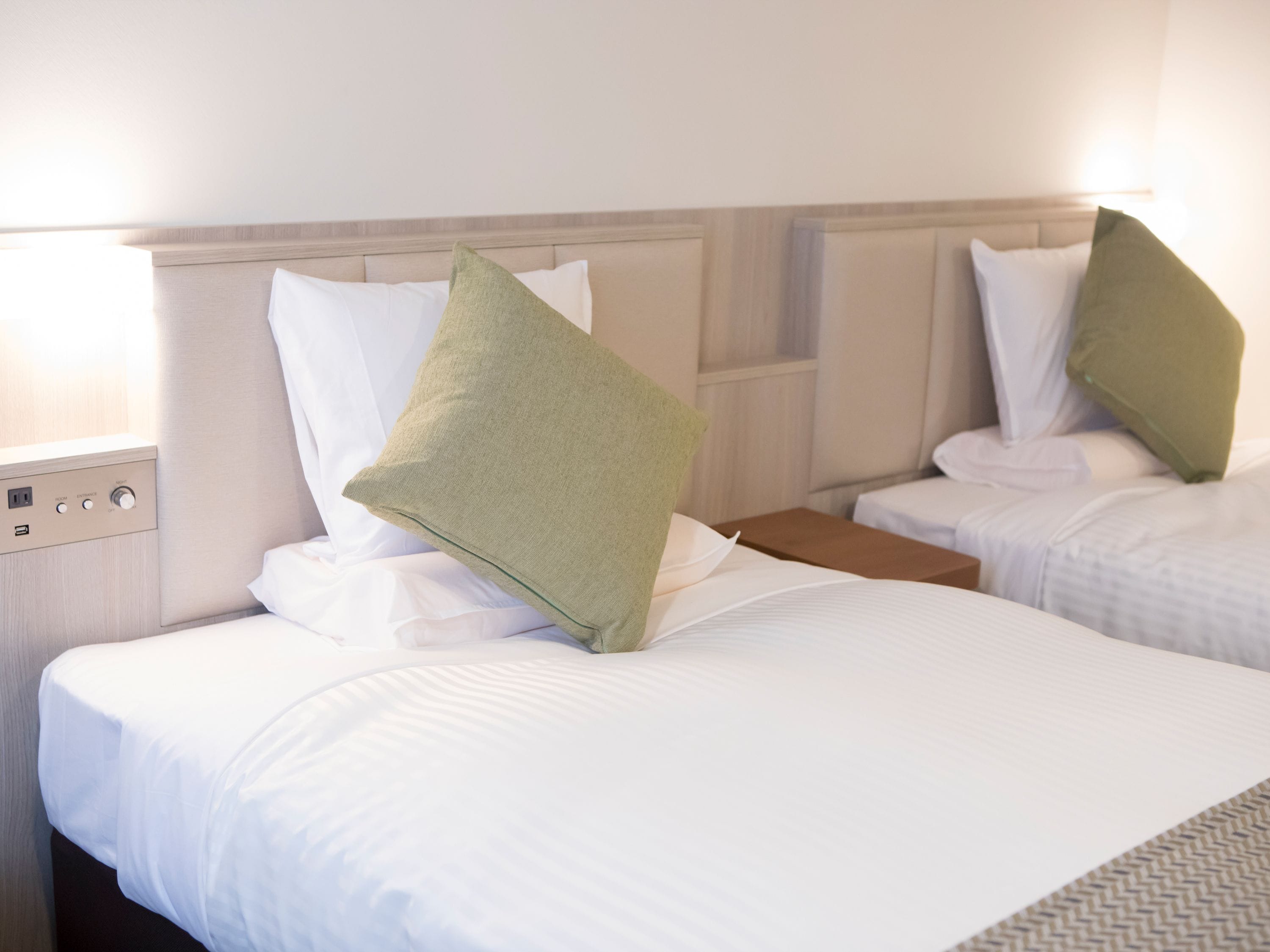 Une chambre d’hôtel avec deux lits, chacun doté de draps blancs et d’une tête de lit en bois. Une table de chevet avec une lampe et une prise électrique se trouve sur le côté gauche de chaque lit.