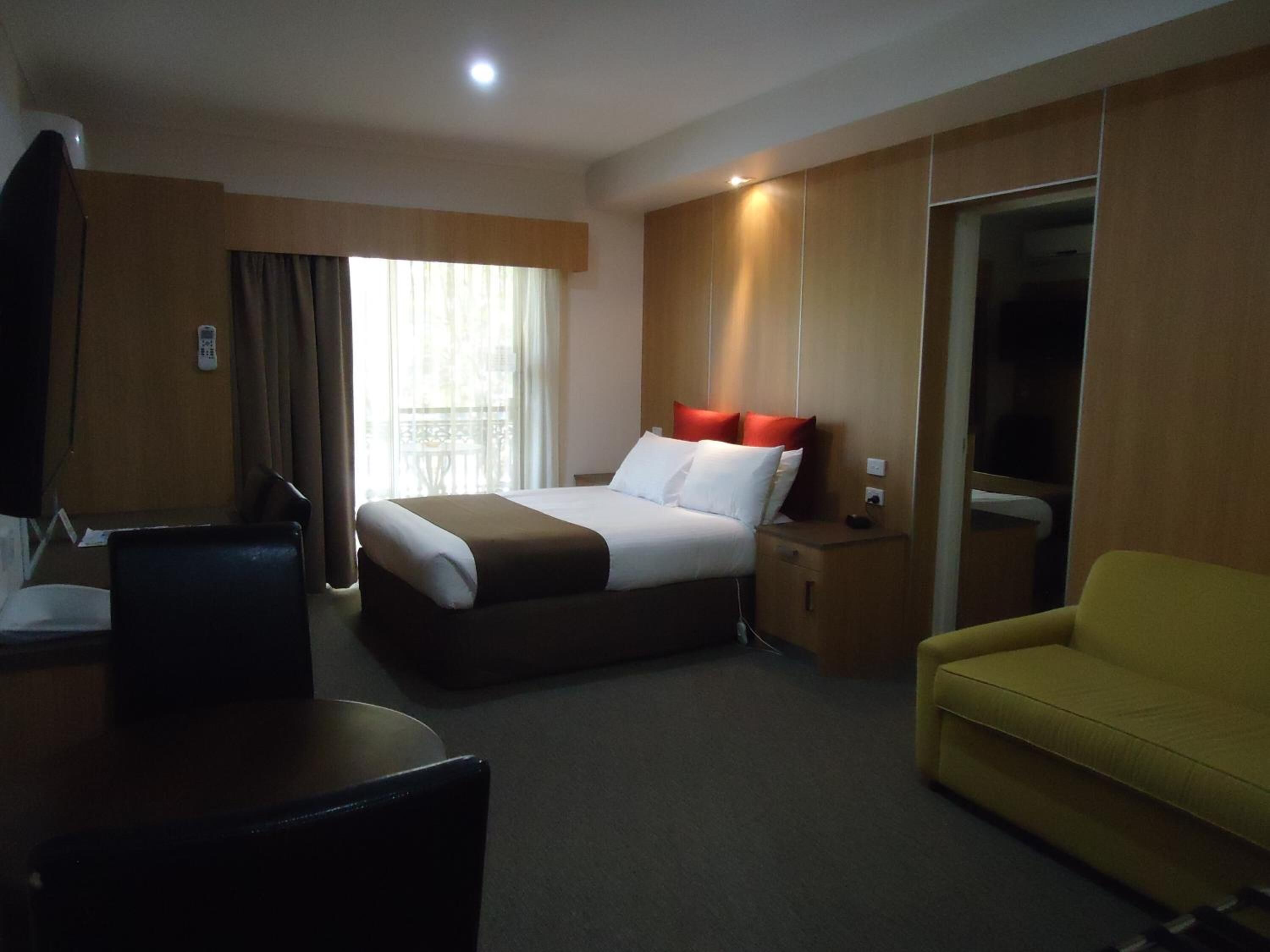 A hotel room with a bed, a yellow sofa, a desk with a chair, a TV, and a window with curtains.