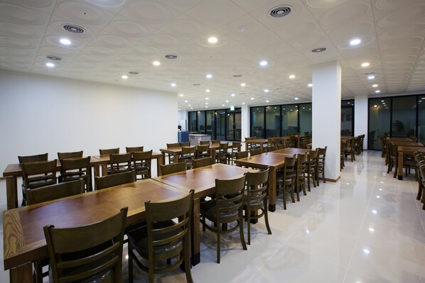 A spacious dining area with multiple wooden tables and chairs, large windows, and a modern ceiling design.