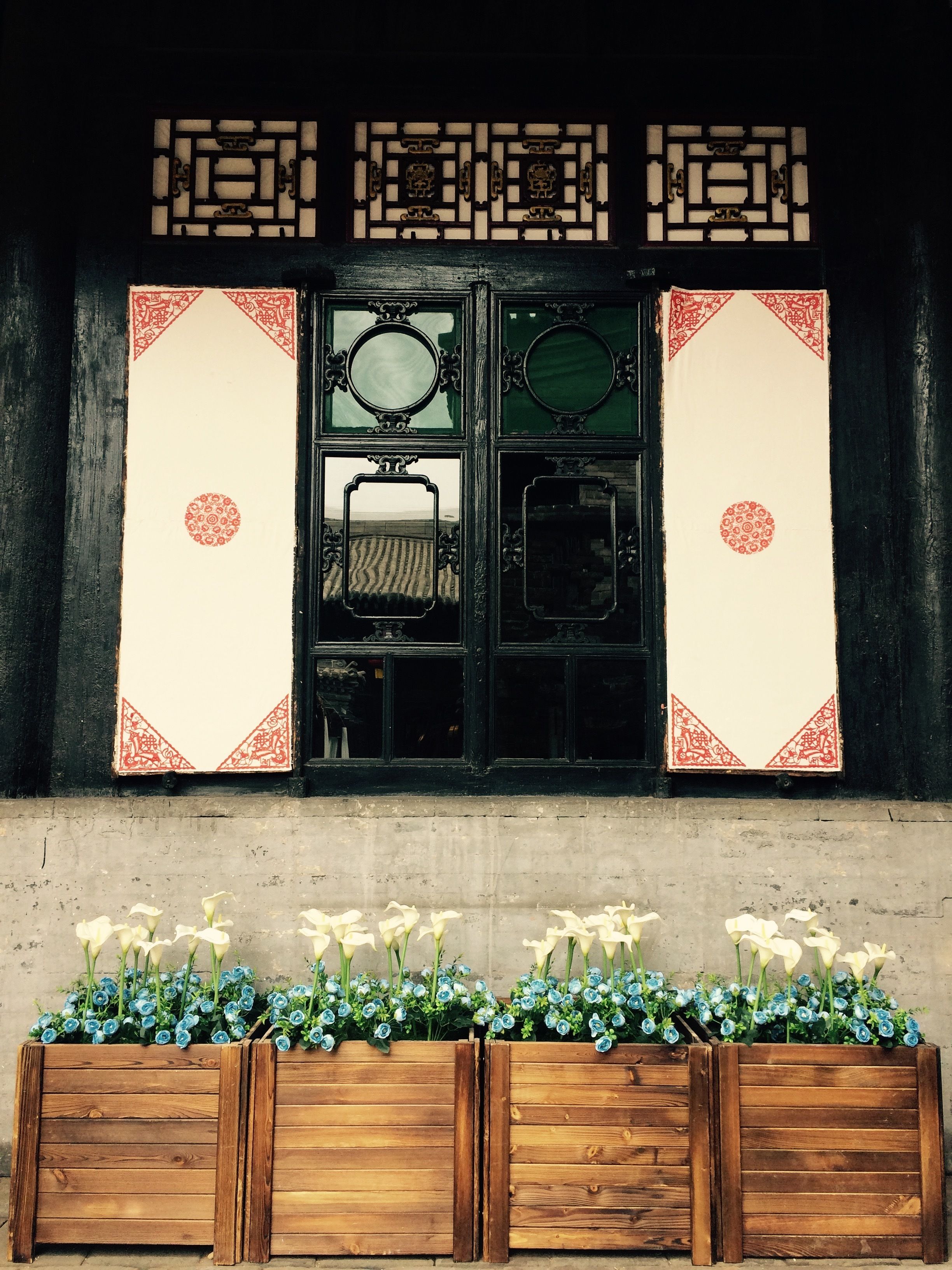 A traditional Chinese window with ornate designs and mirrors. Below, there are wooden planters with white and blue flowers.