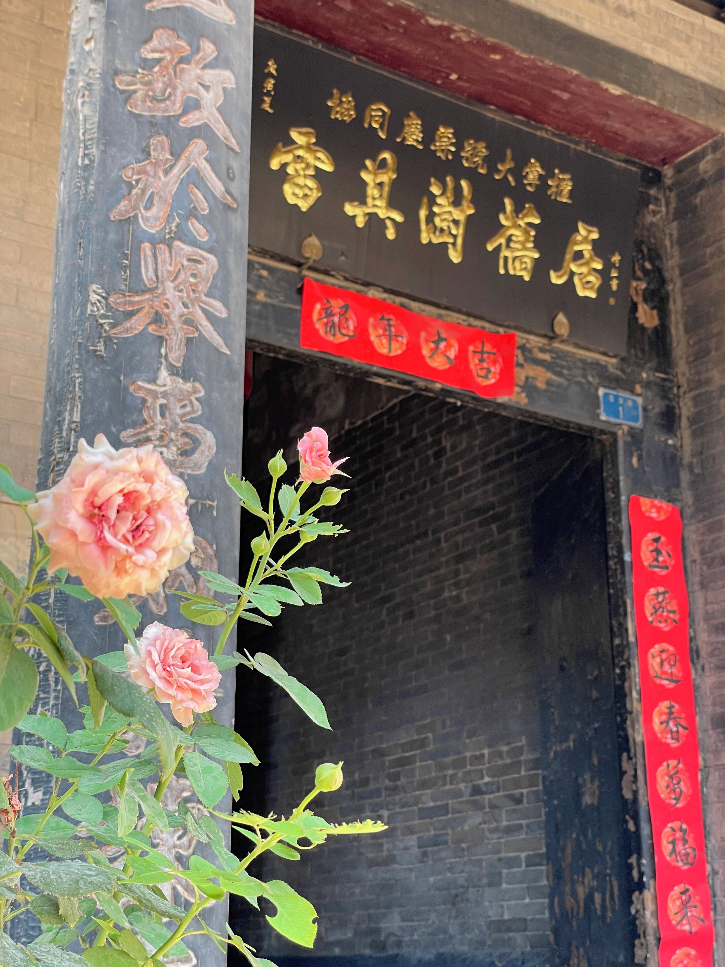 A building entrance with a sign in Chinese characters, a red banner with Chinese characters, and a rose bush in the foreground.