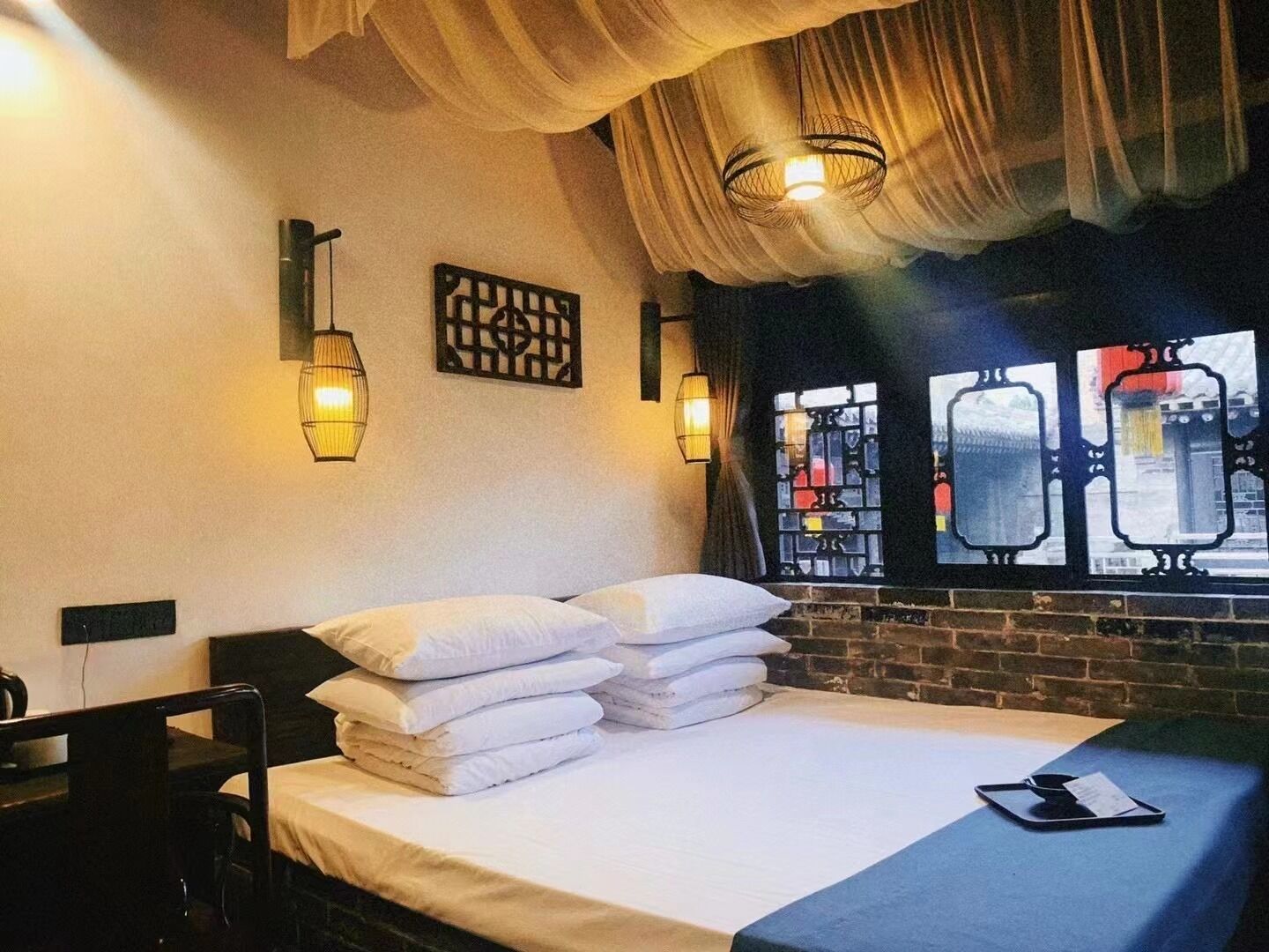 A hotel room with a bed, pillows, a bedside table, a chair, a window with a view, and wall-mounted lights.