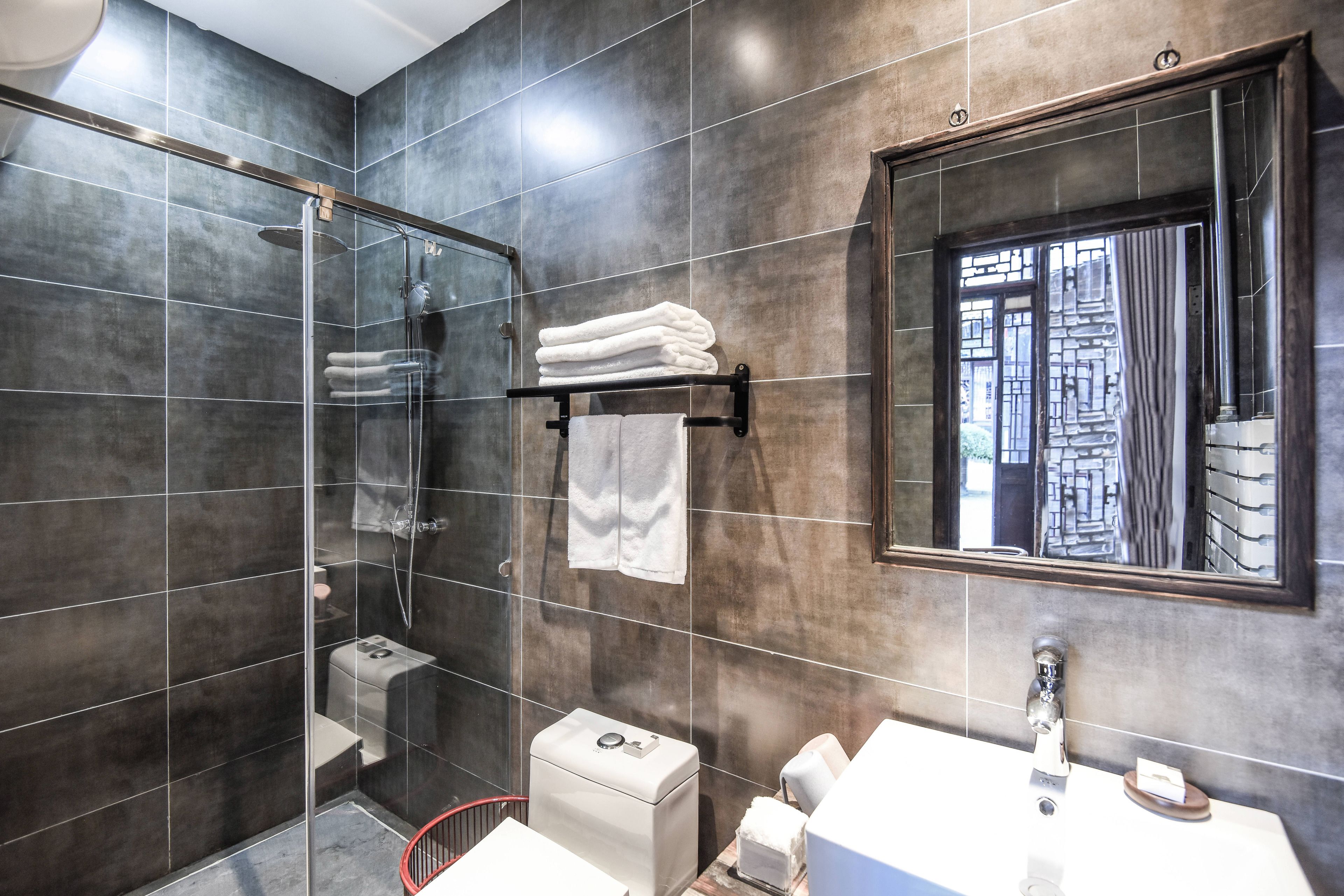 A modern bathroom with dark tiles, a glass-enclosed shower, a white sink, and a large mirror.