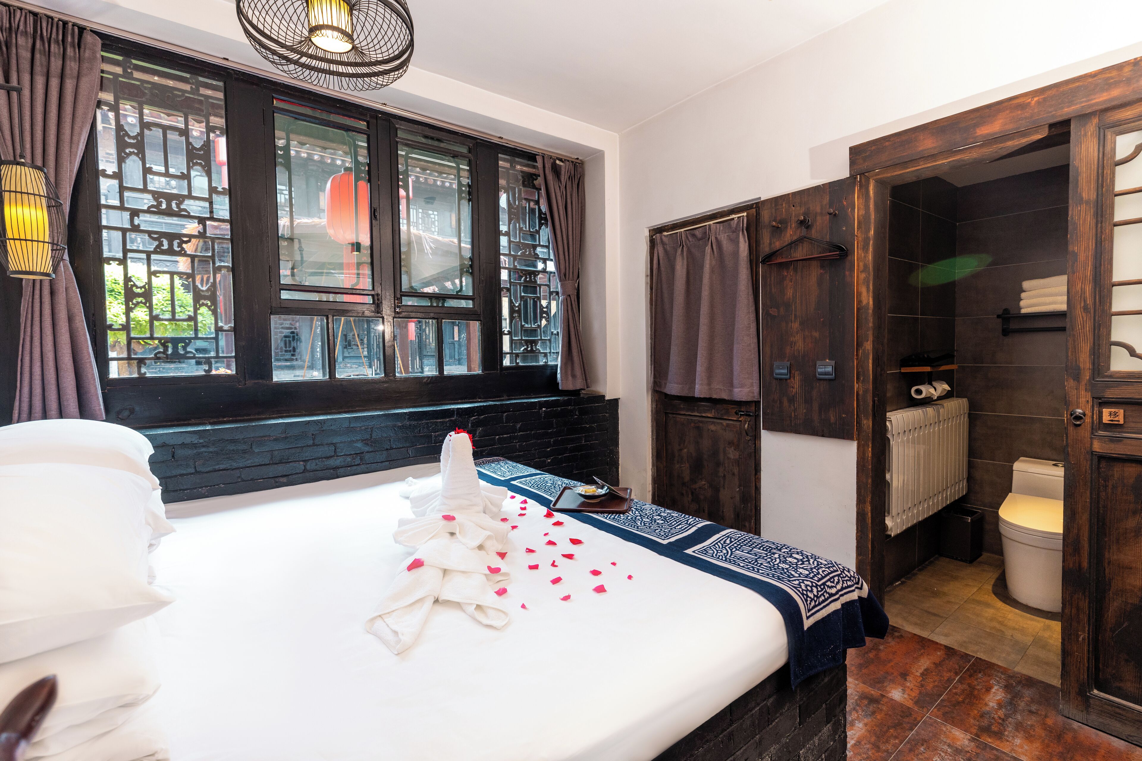 A hotel room with a large bed, a window with curtains, a wooden wardrobe, and a bathroom visible through an open door.