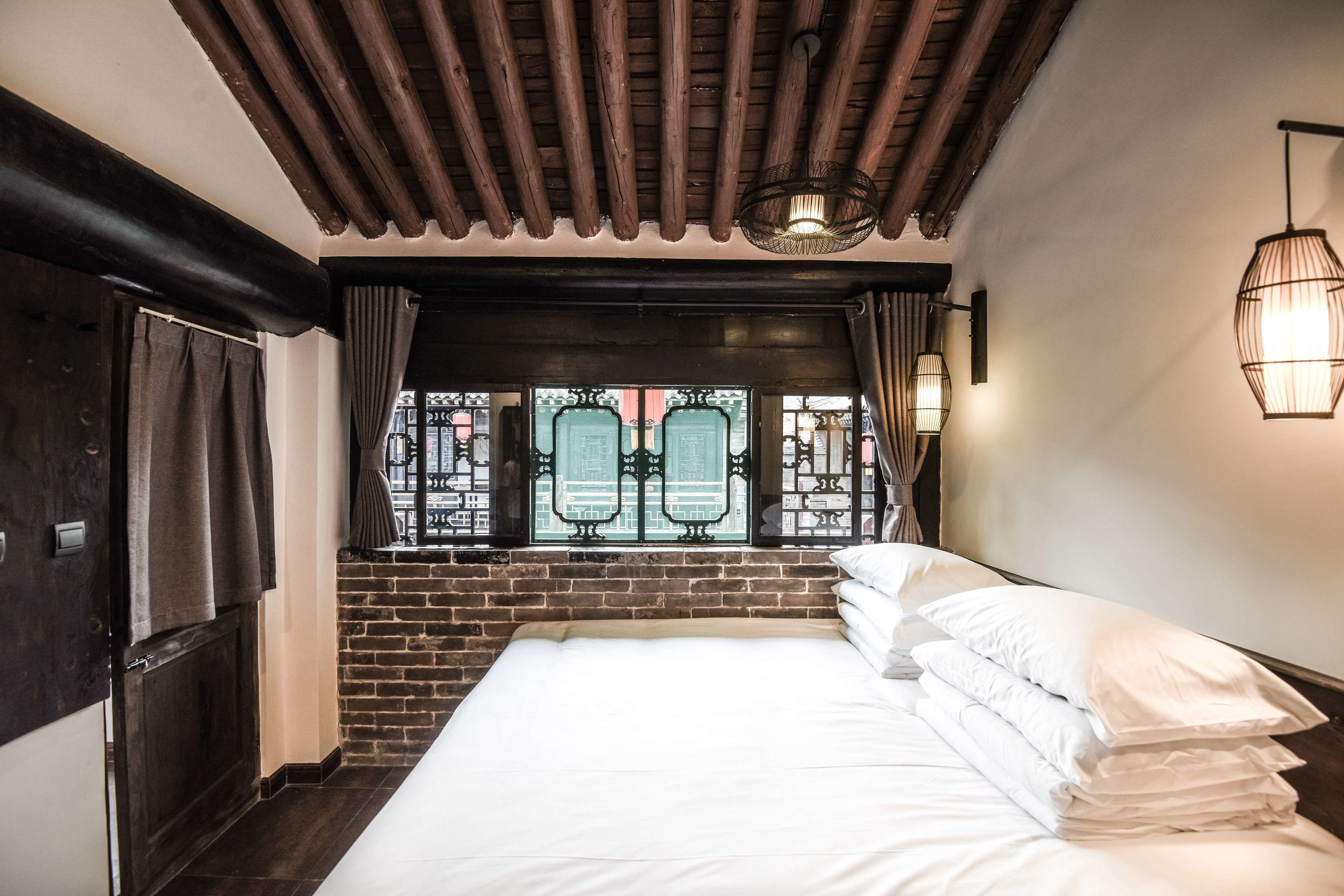 Habitación de hotel con una cama grande, vigas de madera en el techo, pared de ladrillo y vistas a través de una ventana con rejas decorativas.