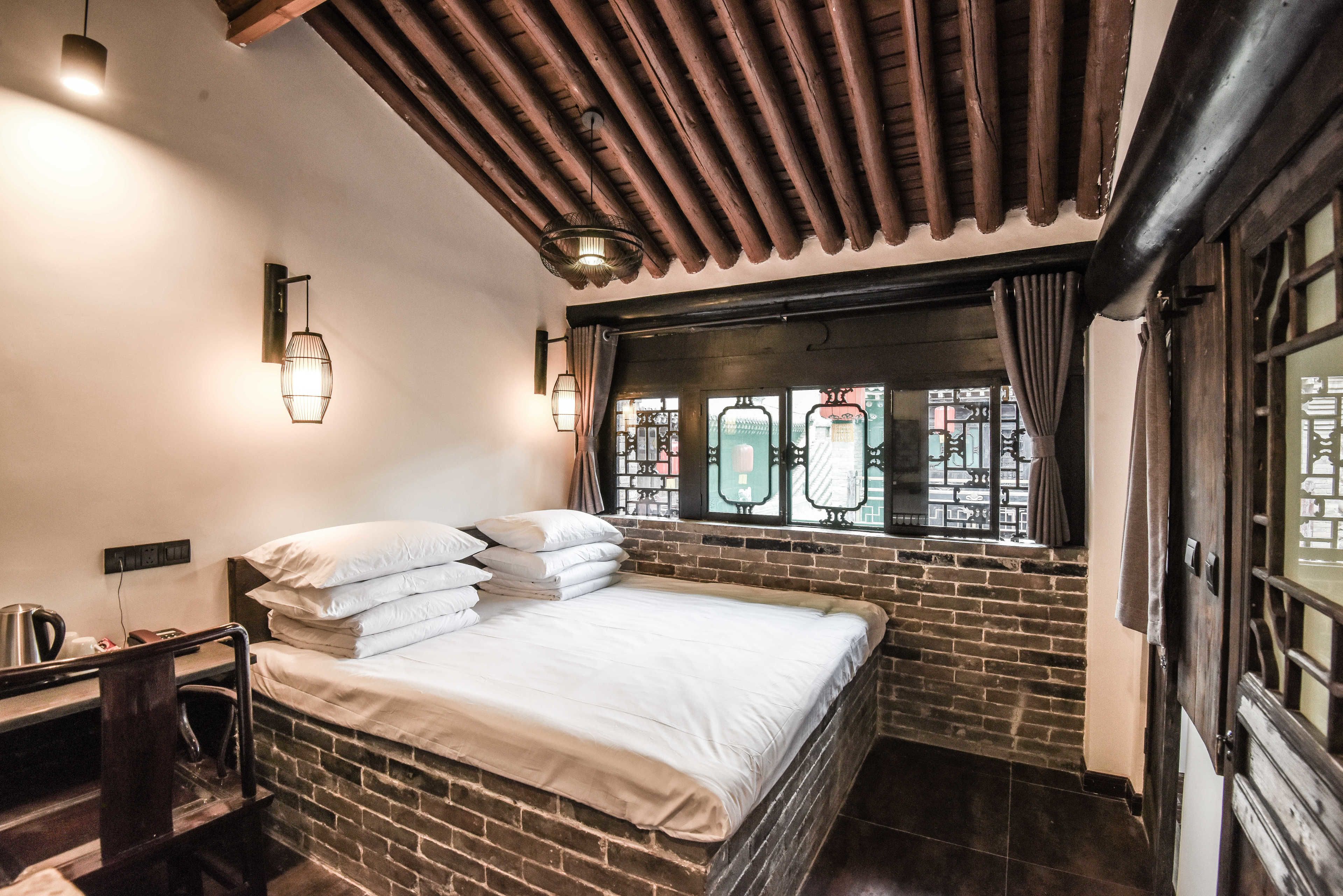 A room with a brick wall, a bed with white linens, a wooden headboard, a nightstand with a kettle, and a window with decorative iron bars.