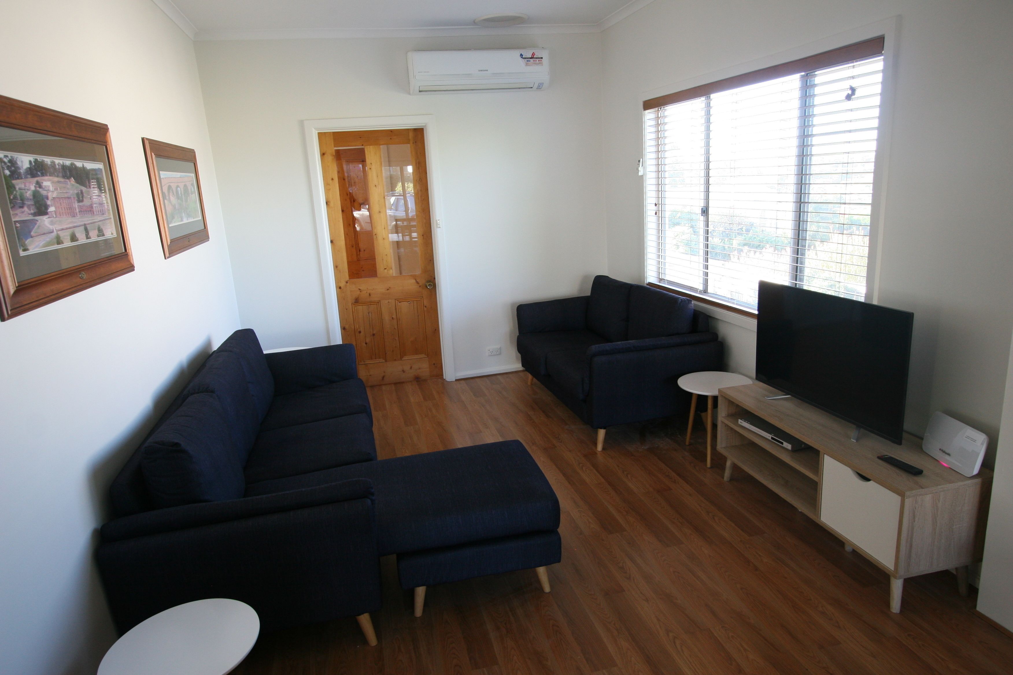 A living room with a dark blue sofa set, a small white table, a TV on a wooden stand, and a door leading to another room.