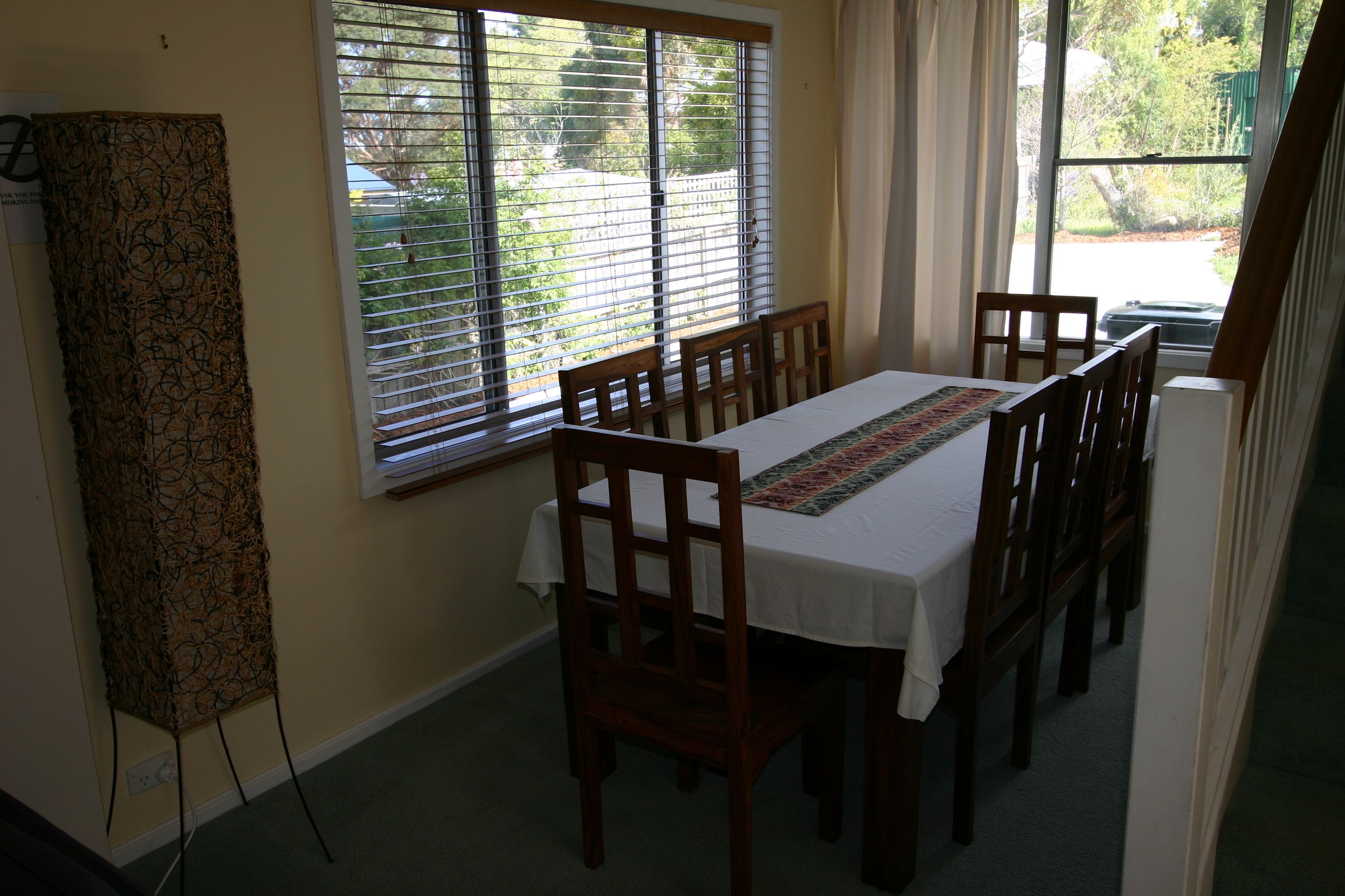Un coin repas avec une table dressée pour six personnes, des chaises en bois et une fenêtre équipée de stores.