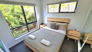 Ein Schlafzimmer mit einem großen Bett, zwei Nachttischen und zwei Fenstern mit Blick auf Grünflächen.