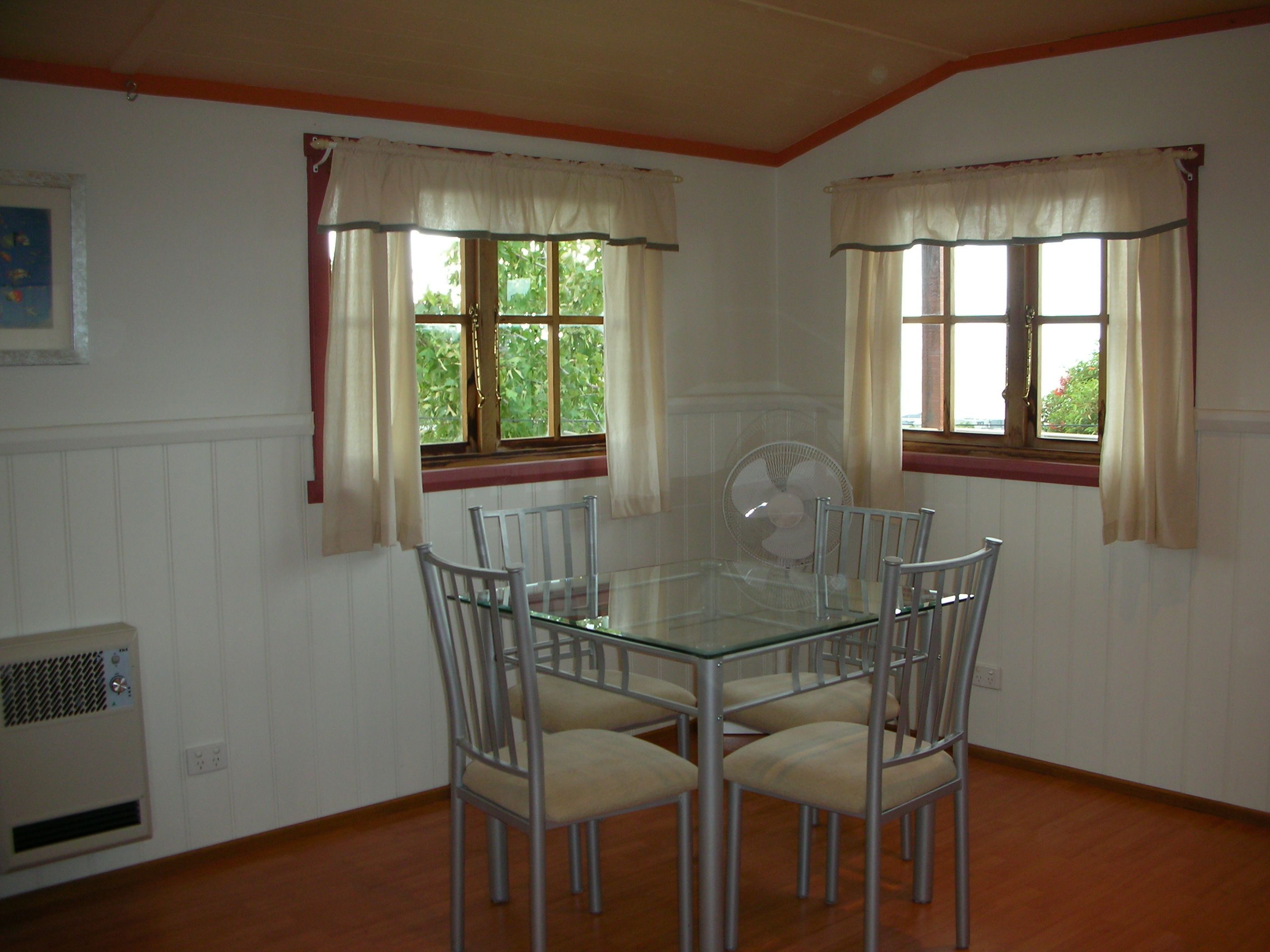 A dining area with a glass table surrounded by four chairs, two windows with curtains, and a wall-mounted fan.