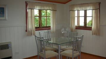 A dining area with a glass table surrounded by four chairs, two windows with curtains, and a wall-mounted fan.