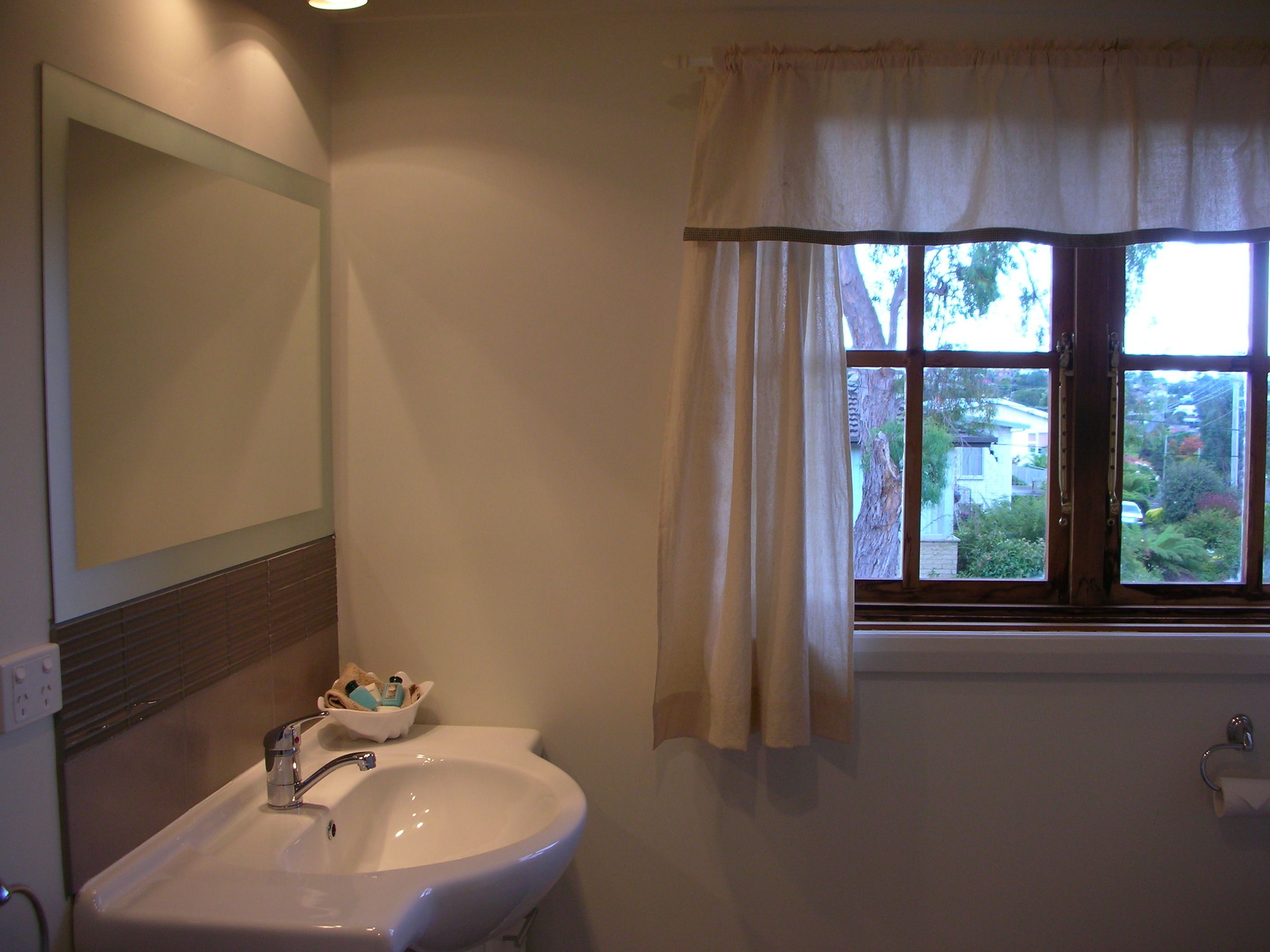 A bathroom with a sink, mirror, window with curtains, and a view of the outside.