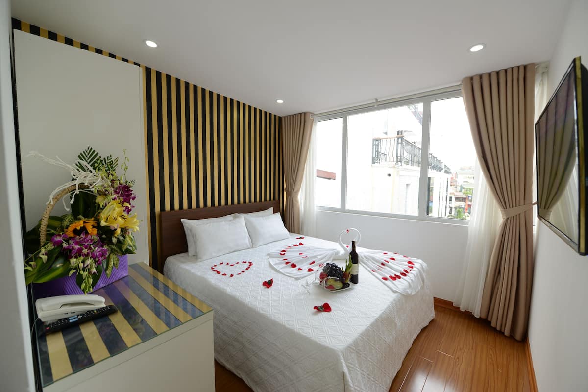 Ein Hotelzimmer mit einem Bett, einem Nachttisch mit Blumen in einer Vase, einer Flasche und einer Schale mit Obst.
