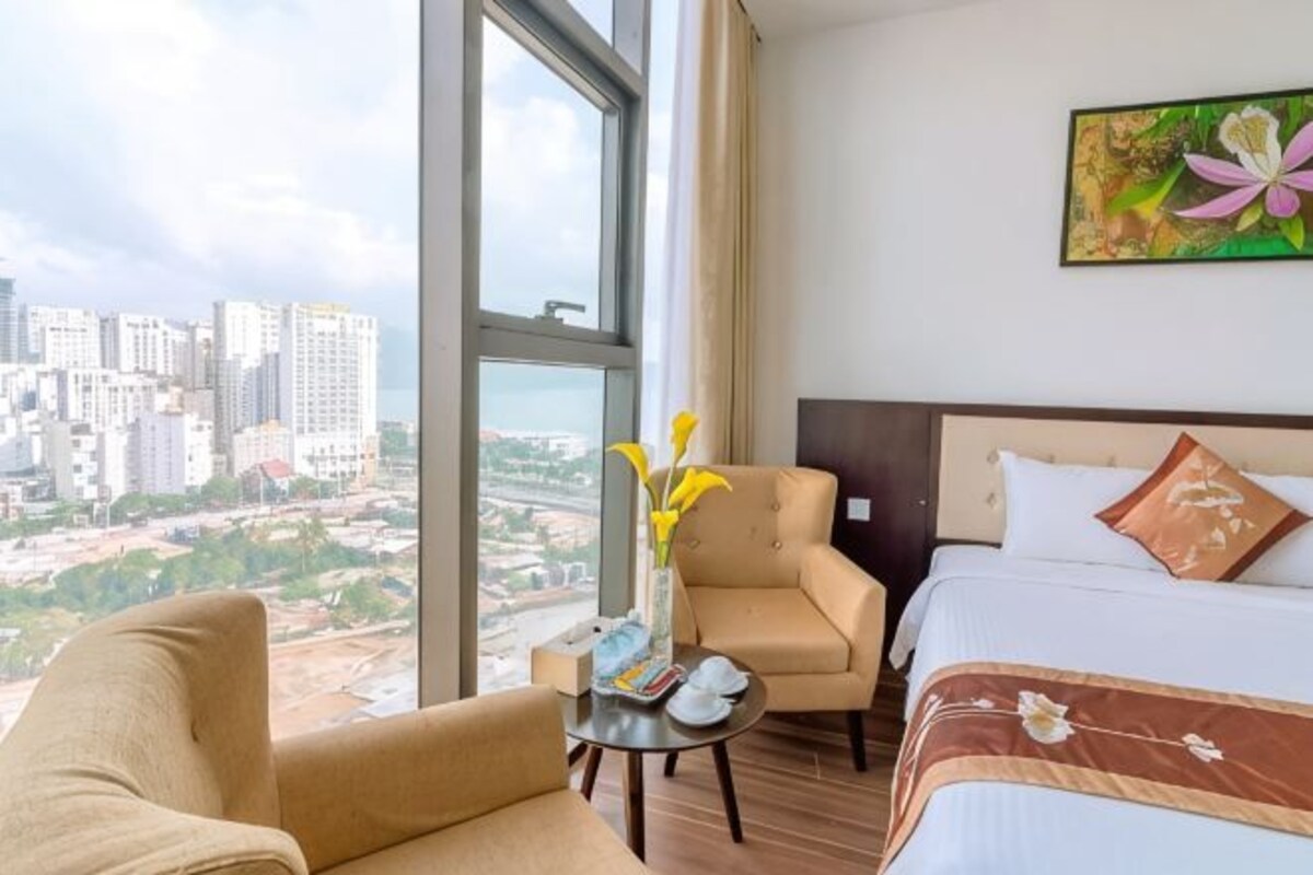 A hotel room with a bed, a chair, a small table, a vase with flowers, and a painting on the wall.