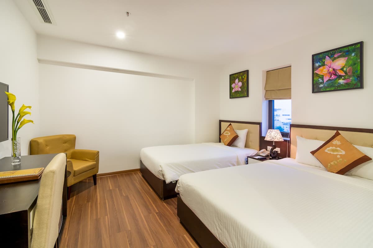 Quarto de hotel com duas camas, uma escrivaninha, uma cadeira, abajur e obras de arte nas paredes.