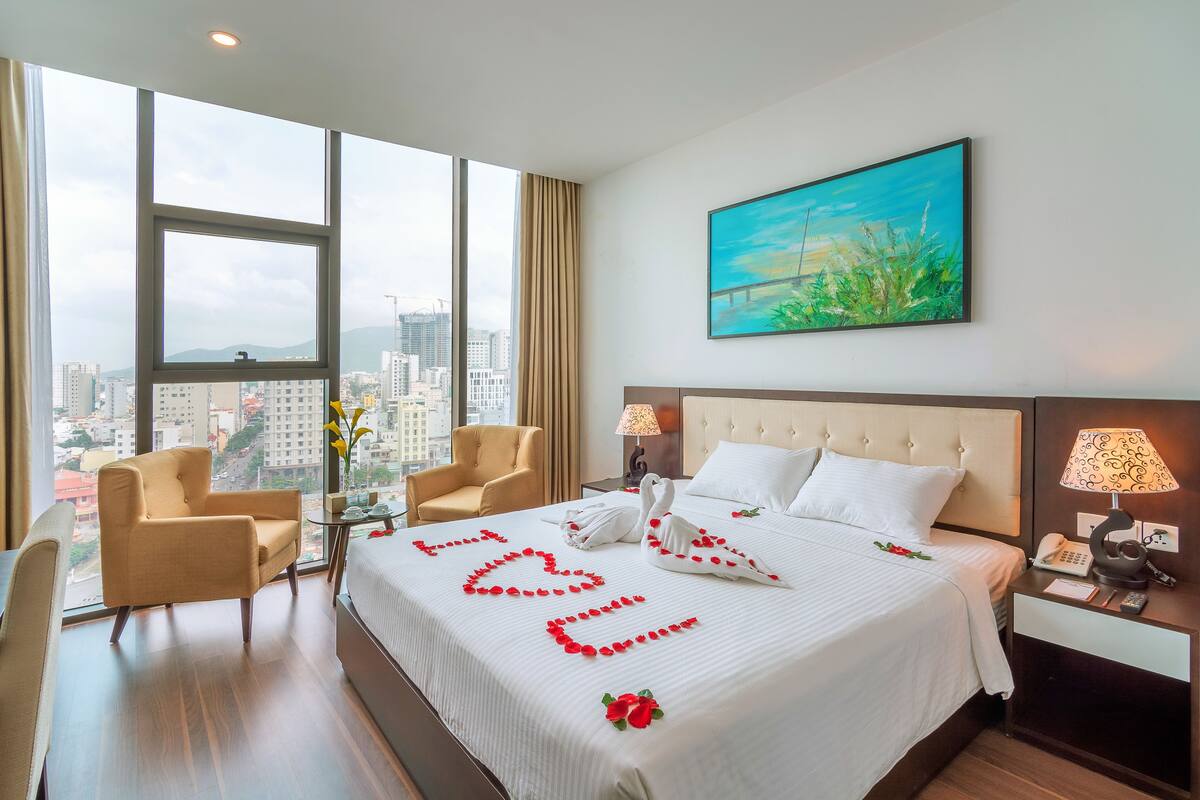 Quarto de hotel com uma cama grande, duas cadeiras, uma mesa de cabeceira e vista para a paisagem urbana.