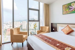 A hotel room with a large bed, two armchairs, a small table with a vase of flowers, and a city view through large windows. - Dana Marina Hotel (Da Nang)