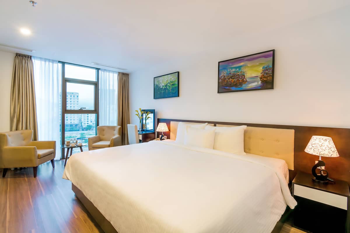 Quarto de hotel com cama grande, duas poltronas, mesa de cabeceira com luminária, quadro na parede e vista para edifícios pela janela.